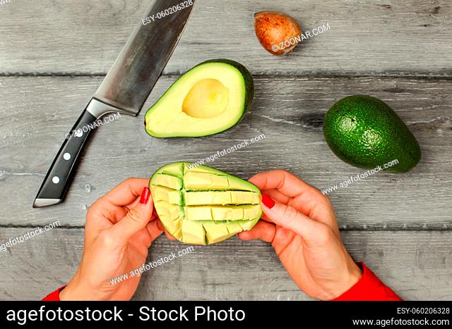 Tabletop view, woman hands holding avocado with cuts, preparing it, removing pulp. Whole avocado, seed and knife on gray wood desk next to it
