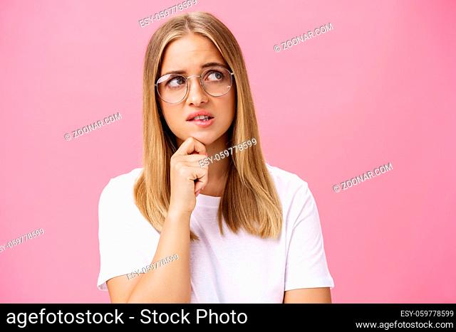 Studio shot of insecure smart nerdy woman in glasses and white t-shirt standing troubled and worried touching chin looking bothered at upper right corner...