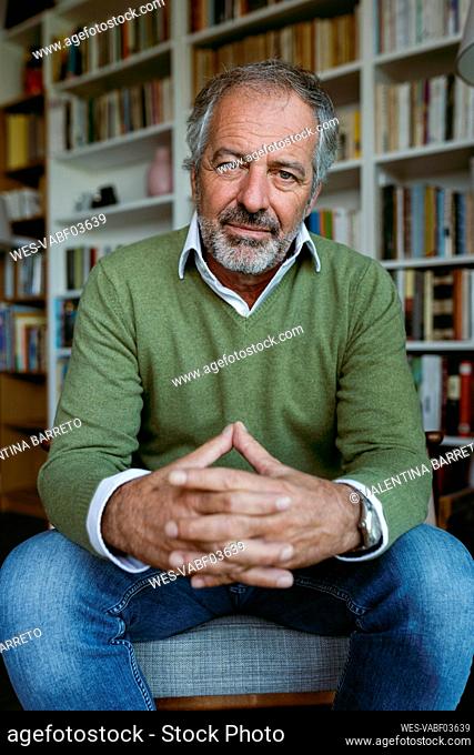 Man with hands clasped sitting on hassock against bookshelf at home
