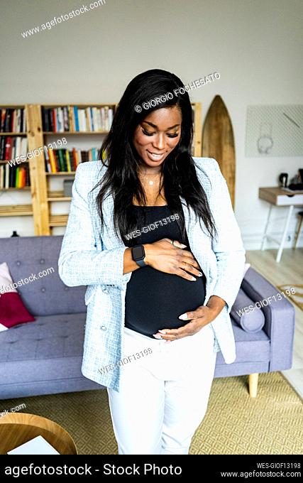 Happy young pregnant female freelancer touching abdomen while standing at home