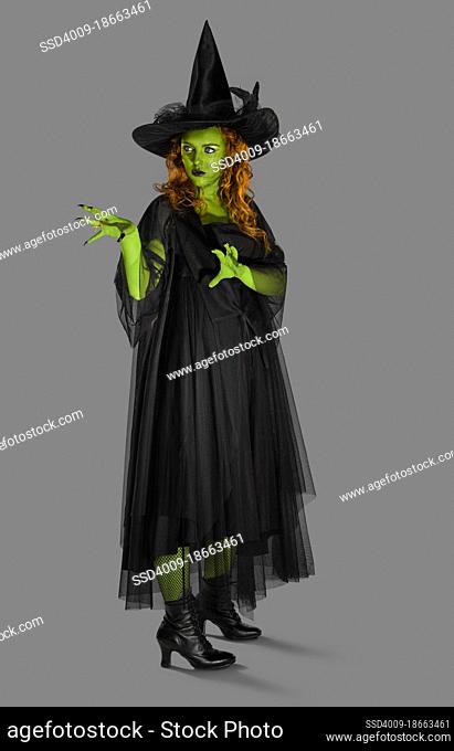 Portrait of a woman dressed as a witch against a gray background with green body paint, looking off camera with her hands up casting a spell