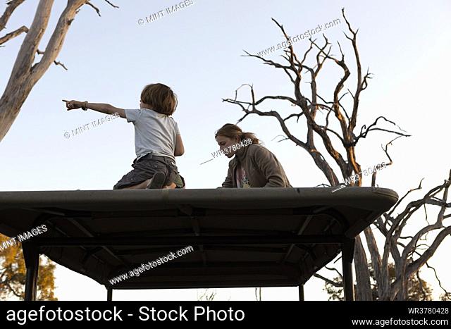 Two children seated on top of a safari tourist jeep, in woodland, at sunset
