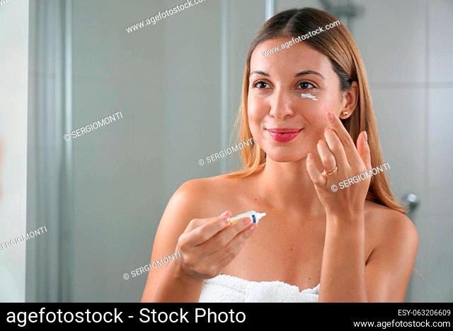 Skin care routine. Portrait of beautiful girl applying cream under eye with finger. Dark circles and anti-wrinkle concept