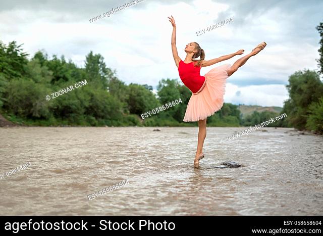 Stylish ballerina stands on the right toe with outstretched arms in the shallow river on the background of green shore and cloudy sky