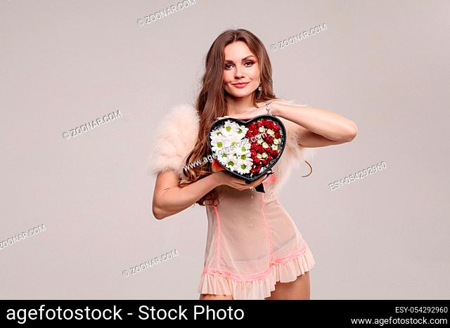 Front view of sexy lady wearing nighty looking at camera and showing present in studio. Attractive woman keeping heart with flowers and posing on grey isolated...
