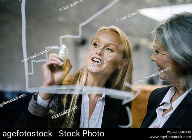 Happy mature and young businesswoman drawing chart on glass pane in office