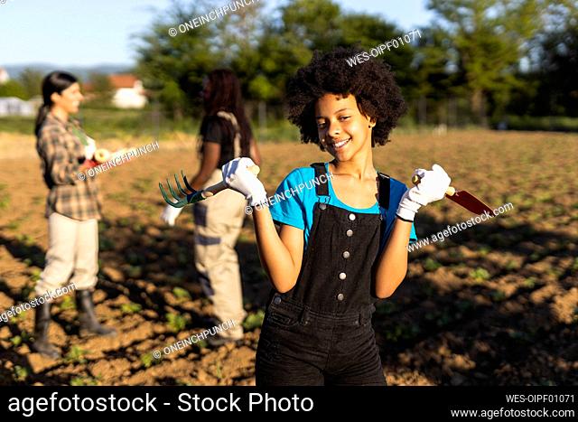 Smiling girl standing with trowel and rake at organic farm during sunny day