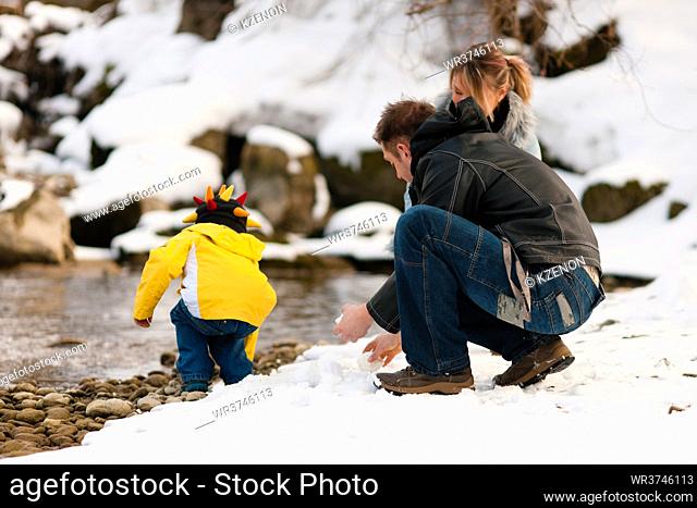 Family - father, mother and son to be seen - on a walk along a riverbank in winter; they are making snowballs