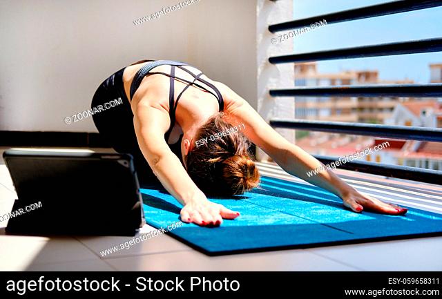 Woman perform yoga child pose on mat use on-line session on tablet. Workout at home in terrace due to self-isolation pandemic corona virus covid-19