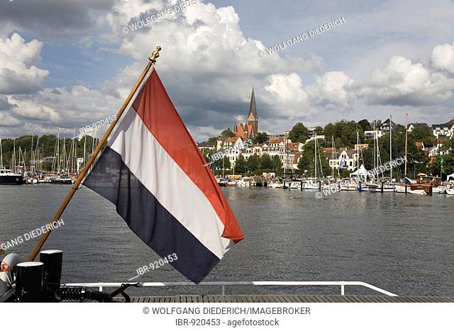 Flag of the Netherlands flying in front of the eastern shore of the Innenfoerde at the sail boat harbour and the Church of St. Juergen or St