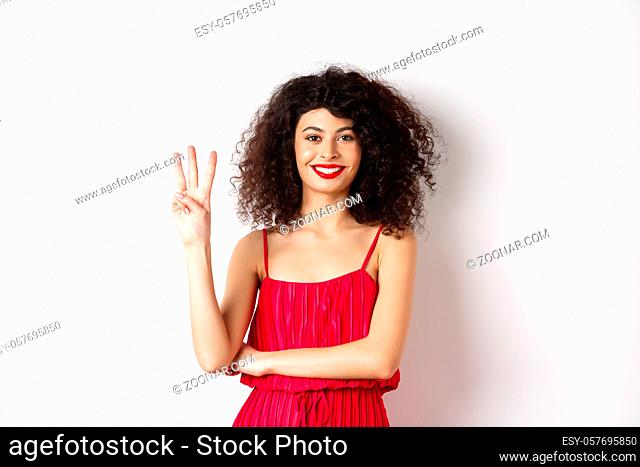Beautiful lady with curly hair, wearing elegant red dress and showing number three, making order, standing over white background