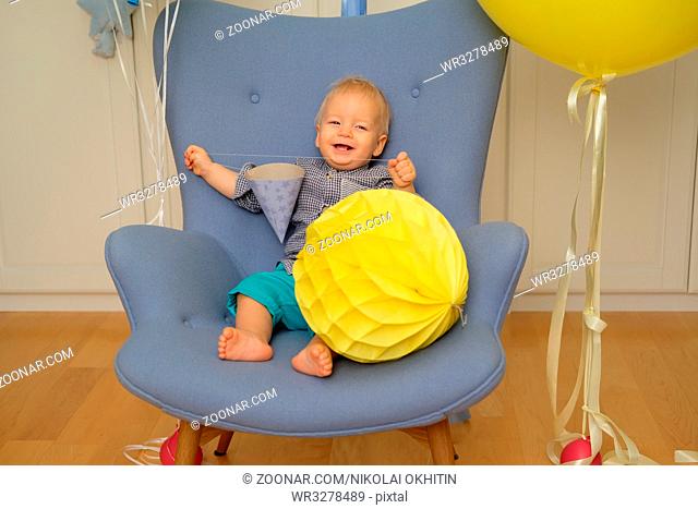 One year old baby boy first birthday. Toddler child sitting in chair and having fun with balloons