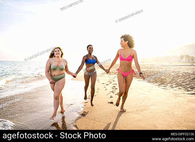 Happy friends holding hands and running at beach on sunny day