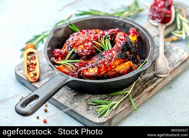 Cast iron pan with baked pork ribs, rosemary, spoon with cherry sauce on wooden serving board, selective focus