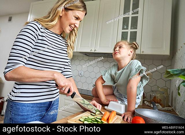 Smiling mother looking at daughter while chopping cucumber in kitchen