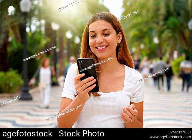 Excited young woman watching her smartphone when walking in the street with blurred people on the background. Millennial girl using mobile app outdoors