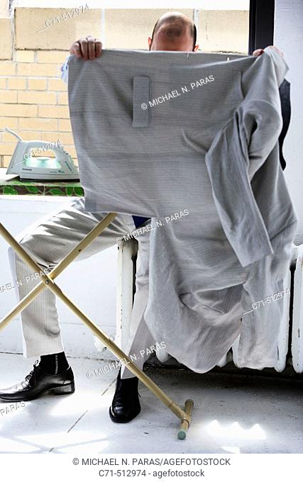 Man sitiing on radiator ironing clothes and hiding face with jacket