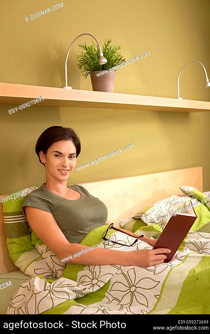 Portrait of mid-adult woman sitting with book in bed, looking at camera, smiling