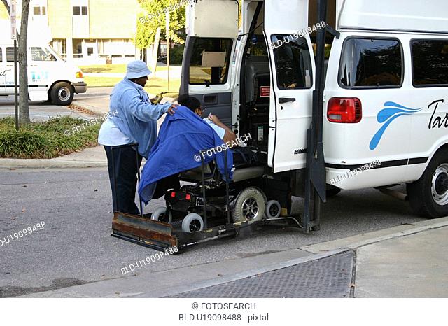 Person in a wheelchair receiving assistance in using a lift as he prepares to get into a van