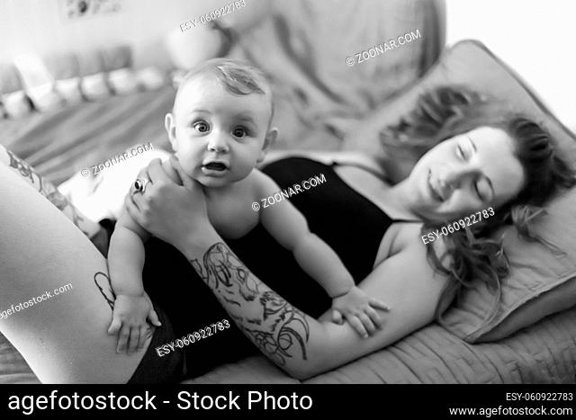 Mother and baby son joyful snuggles. Baby sitting on his mother's belly and looking at camera with a surprised expression. Black and white portrait