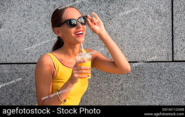 Young attractive female fashion model wearing fashionable yellow dress and drinking orange cocktail. Smiling woman wearing sunglasses