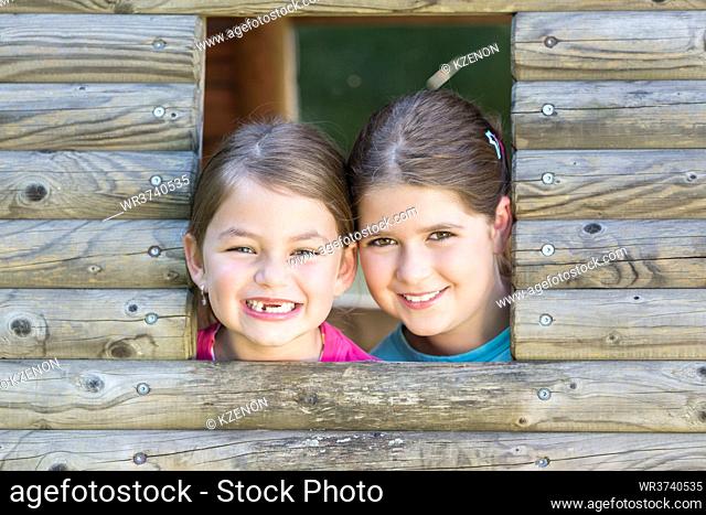 Two little girls looking out of playhouse window on playground and grin