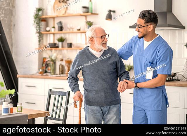 Smiling friendly in-home male nurse in uniform supporting an old man with a walking stick