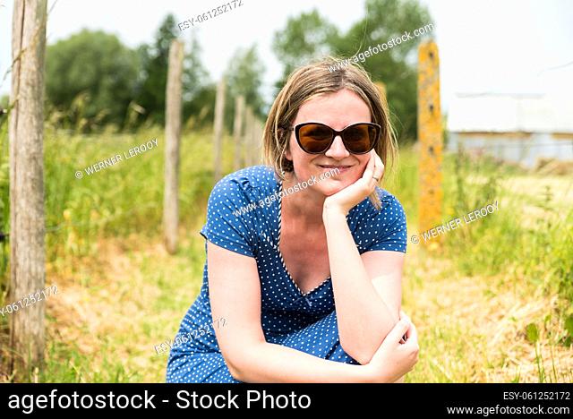 Portrait of an attractive thirty year old girl with a blue dress and sunglasses sitting at the Flemish countryside, Belgium