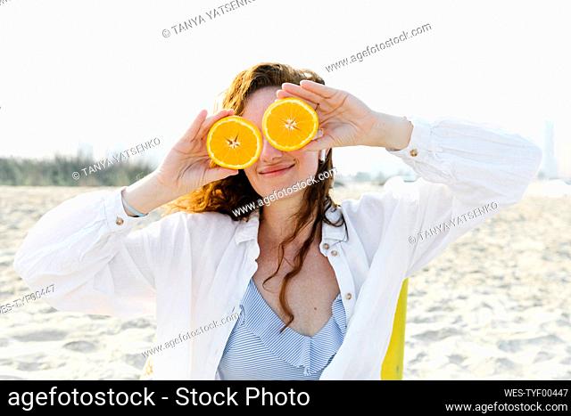 Smiling woman covering eyes with oranges at beach on sunny day