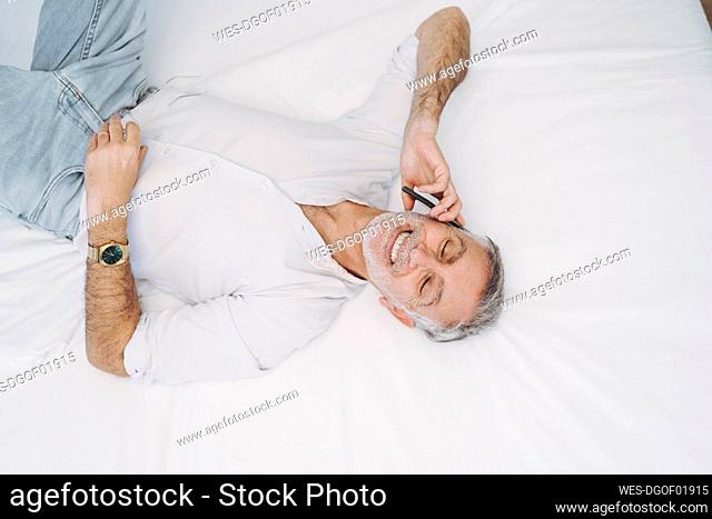 Smiling man talking on smart phone while lying on bed