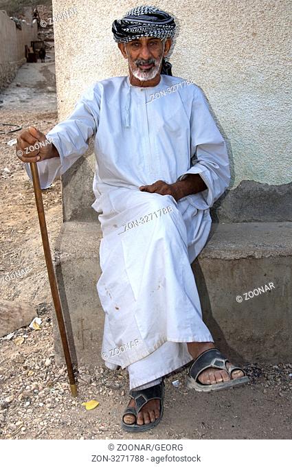 Alter Omani Mann mit Gehstock sitzt vor seinem Haus und blickt in die Kamera, Ash Shab, Sultanat Oman / Old Omani man with a cane sitting on ledge in front of...