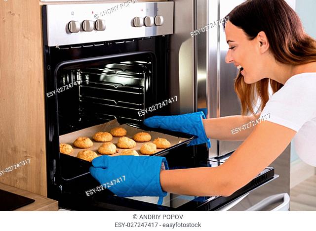 Young Happy Woman Placing Tray Full Of Cookies In Oven At Home