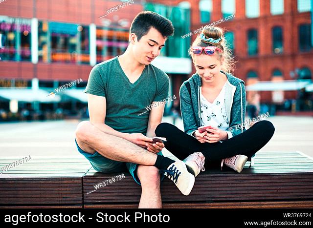 Couple of friends, teenage girl and boy, having fun together with smartphones, sitting in center of town, spending time together