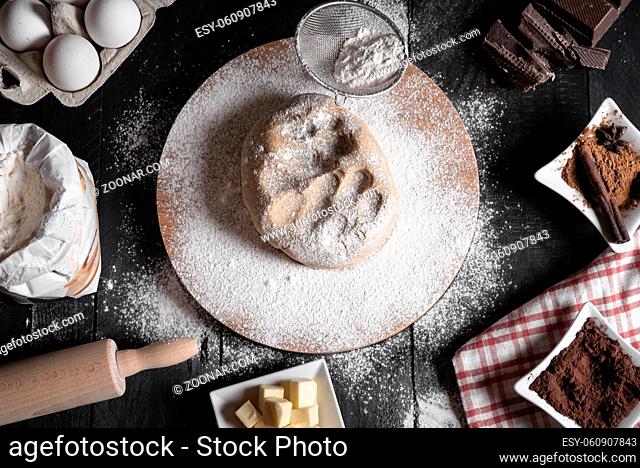 Christmas cookies dough on a wooden platter, surrounded by flour, eggs, cinnamon, chocolate, butter, and a rolling pin, on a rustic black table