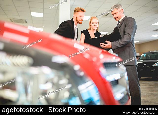 Vehicle dealer selling electro car to young couple. Man and woman talking with sales manager in auto showroom