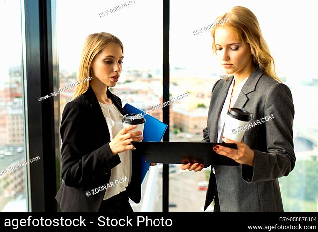 Business women discuss documents and drink coffe in office in front of panoramic windows with view at city