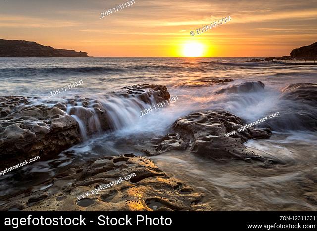 Sun rising on the horizon shining its golden light down Long Bay to Malabar with rock flows in the foreground