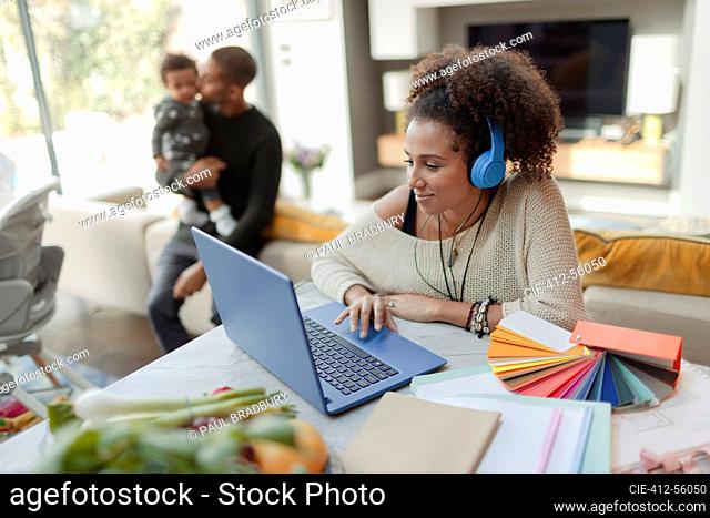 Woman working at laptop with husband and baby daughter in background