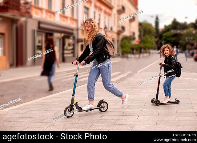 Side view of a young mom and daughter are riding scooters in the city. They have curly hair and the same clothes