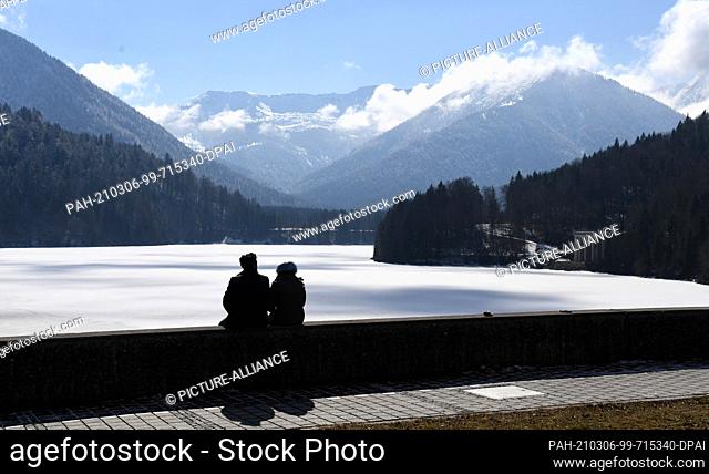 06 March 2021, Bavaria, Lenggries: A woman and a man sit on the dam wall of the Sylvenstein reservoir and look out over the alpine scenery with Mount Lerchkogel