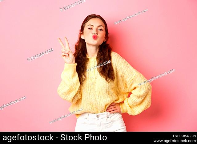 Young glamour woman pucker her lips, making kissing face and v-sign, show peace gesture, standing against pink background