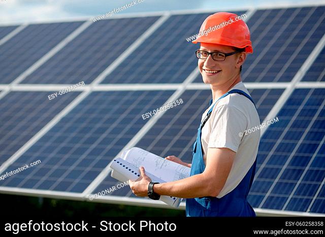 Young man working with solar panels. Worker at solar power station holding documents looking in camera and smiling