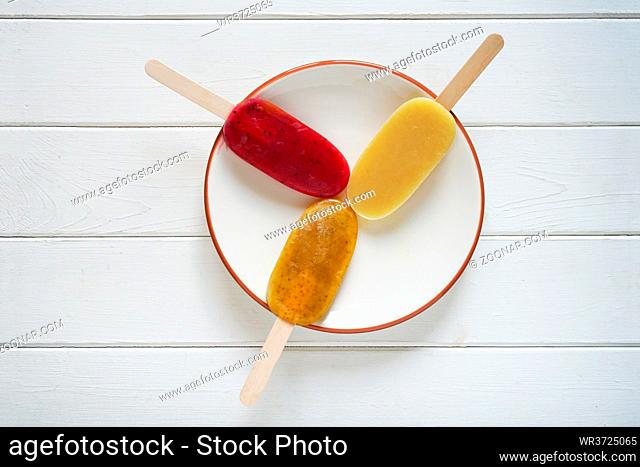 overhead view of three different fruit smoothie popsicles or ice pops on a plate on white wooden background