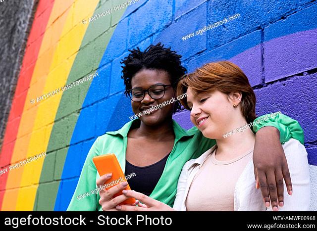 Smiling woman sharing smart phone with friend in front of rainbow colored wall