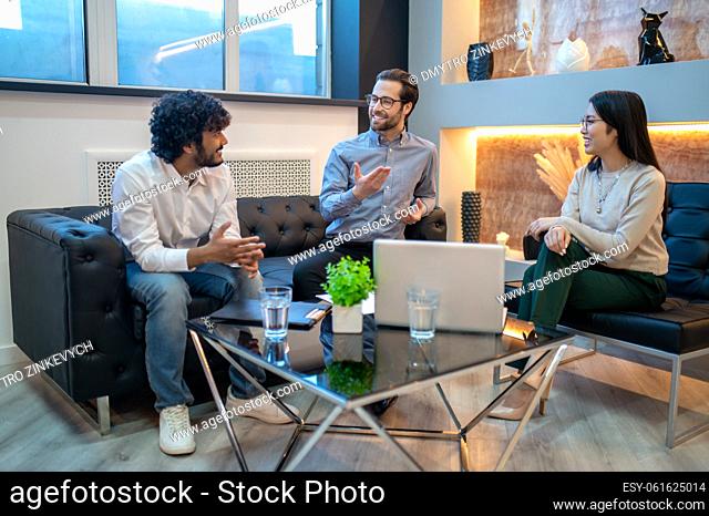 Smiling cheerful businessman seated around the table with his business partners talking to one on them