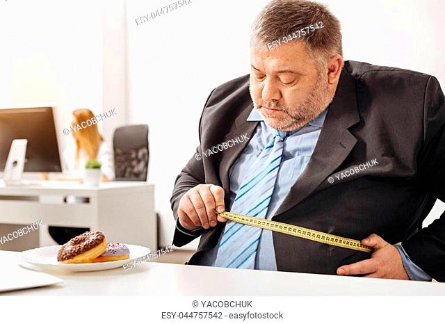 To eat or not to eat. Corpulent hungry indecisive man making some measurements and thinking about changing his lifestyle while a plate with doughnuts standing...