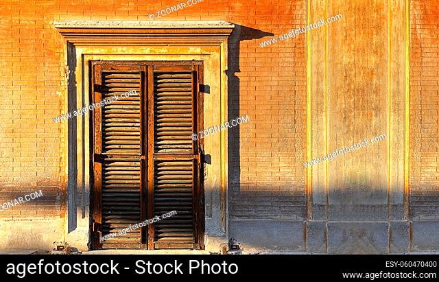 Ancient window with closed wooden shutters illuminated by the sunlight at sunset in Italy. Residential building. Cityscape