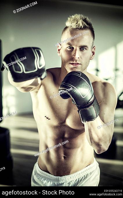 Shirtless handsome muscular young man in gym giving punch towards camera, wearing boxing gloves