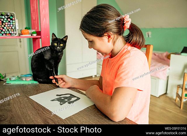 Girl giving felt tip pen to cat sitting on table at home