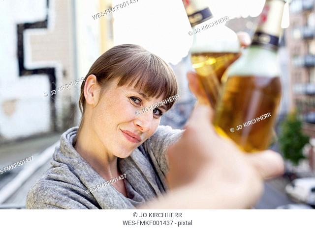 Smiling woman on balcony clinking beer bottle
